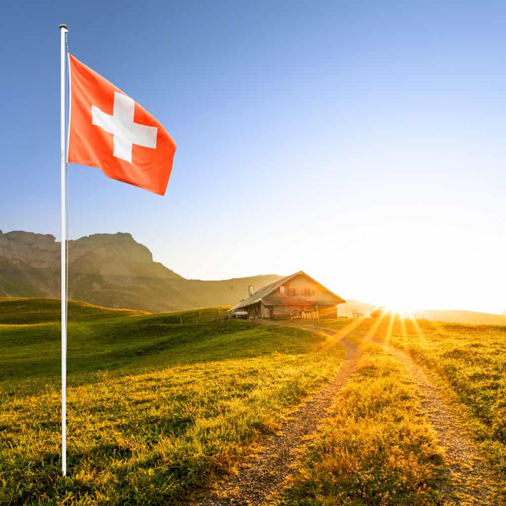 Bergbauernhof bei Sonnenaufgang mit wehender Schweizer Flagge im Vordergrund.