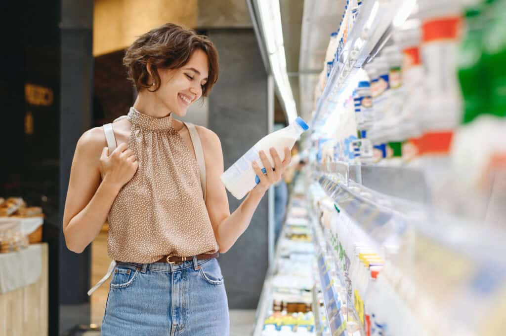 Young smiling happy woman 20s in casual clothes backpack shopping at supermaket grocery store buy choose dairy produce take milk read shelf life inside hypermarket Purchasing gastronomy food concept.
